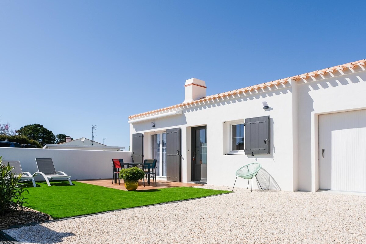 The exterior of a newly built house features a well-maintained garden with synthetic grass and a gravel area. A wooden terrace with a table and chairs is accompanied by two lounge chairs. The bright blue sky enhances the serene atmosphere of the setting.