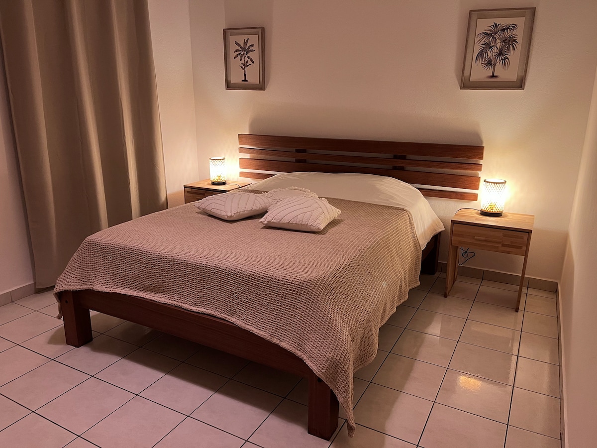 A well-lit bedroom features a spacious bed with a neutral-toned blanket and decorative pillows. Nightstands with lamps flank the bed, and tropical-themed artwork is displayed on the walls. Soft curtains contribute to a calm ambiance in the room.