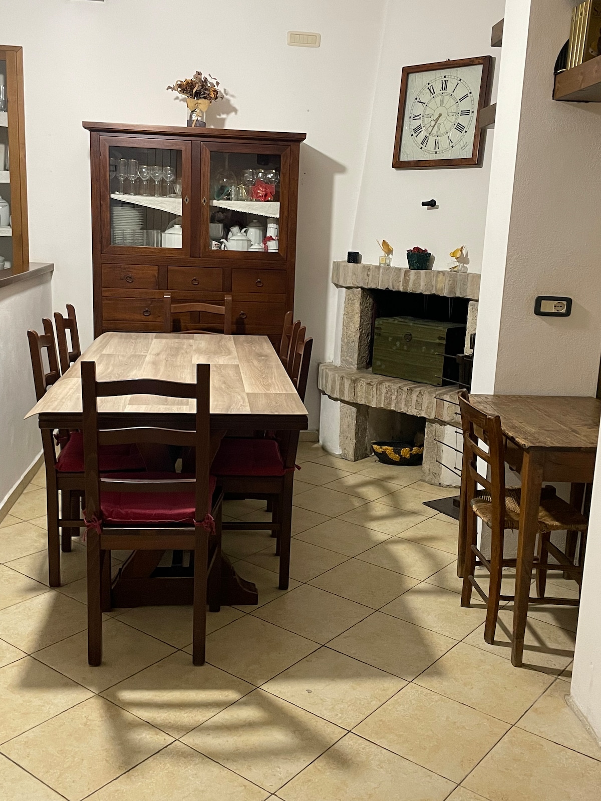 A dining area is presented with a wooden table, surrounded by six chairs with red cushions. A hutch stands against the wall, showcasing glassware, while a stone fireplace adds warmth to the room. Light tiles create a bright atmosphere.