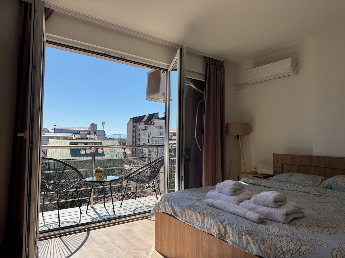 A spacious room is highlighted by a comfortable bed with neatly arranged towels. Large glass doors open to a private balcony with two chairs and a small table, providing a view of the surrounding area under clear blue skies.