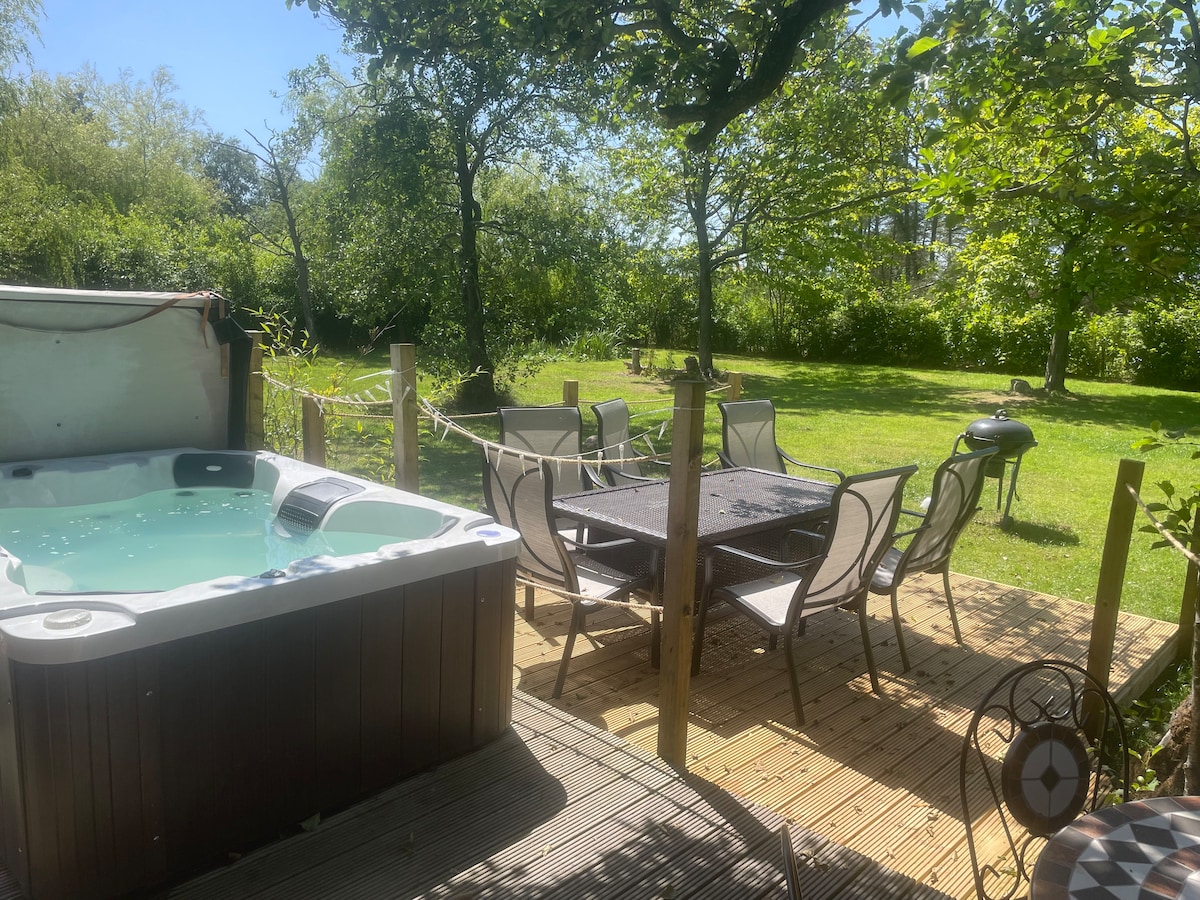 An outdoor seating area features a patio table surrounded by chairs, with a barbecue grill positioned nearby. A hot tub is situated on the deck, and lush greenery and trees create a serene atmosphere in the yard.