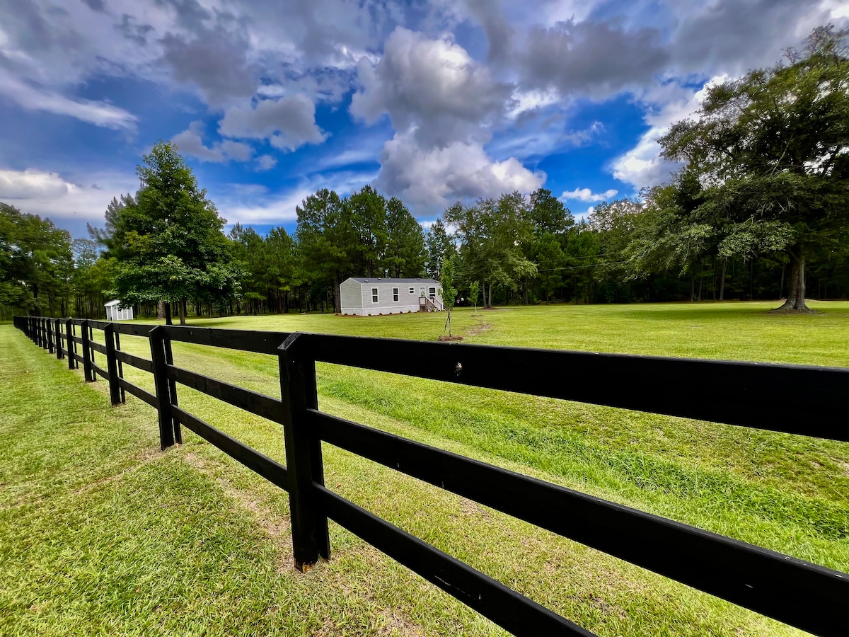 An expansive lawn is framed by a black wooden fence, leading towards a simple white structure nestled among trees. The sky above features a mix of clouds and blue, creating a calm and open atmosphere around the property.