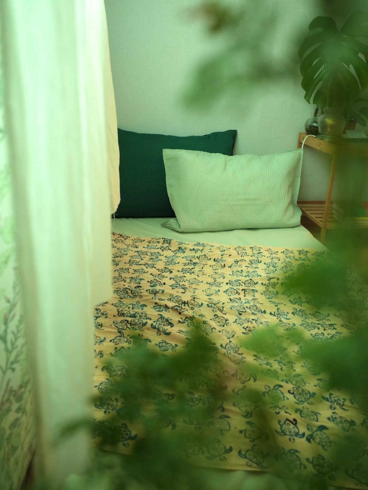 A cozy sleeping nook is depicted, featuring a soft yellow bedding with intricate patterns. Two cushions, one dark green and one light green, are arranged for comfort. The space is framed by soft fabrics and lush greenery, creating a serene environment.