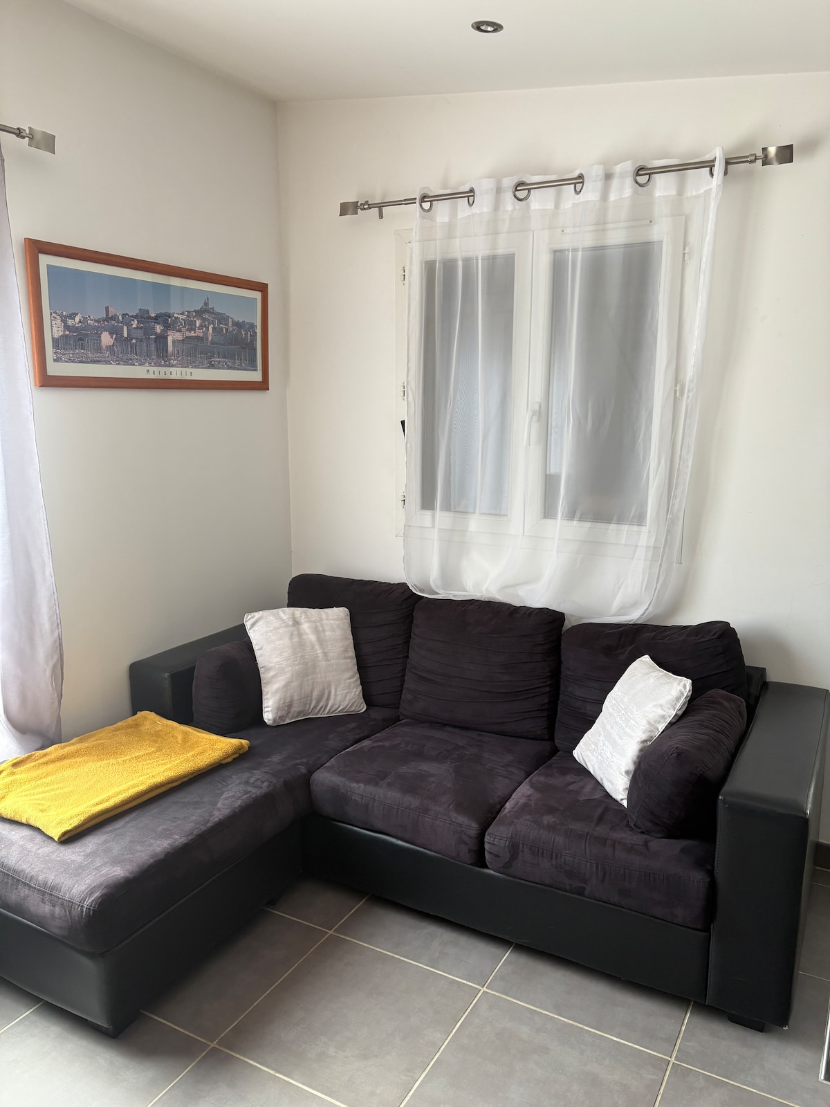 A corner seating area features a dark upholstered sectional sofa adorned with two decorative pillows. A yellow throw provides a pop of color. Natural light enters through a pair of sheer curtains covering the window, and a framed artwork adds interest to the wall.