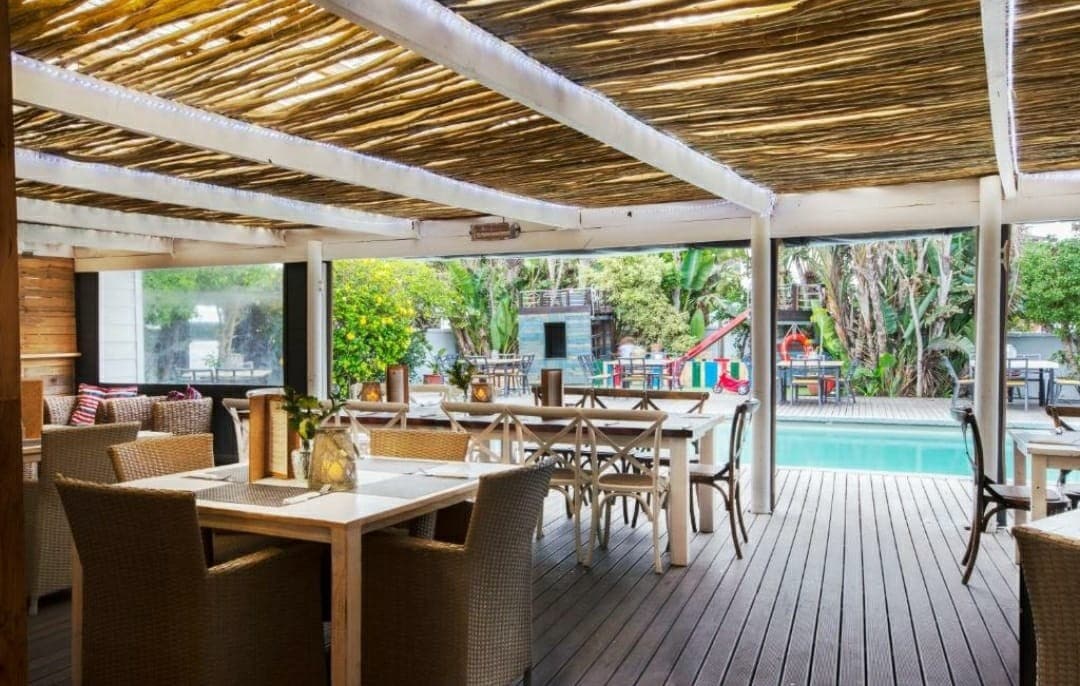 An outdoor dining area features wooden tables and woven chairs beneath a canopy of natural materials. Lush greenery surrounds the space, with a swimming pool visible in the background. Natural light enhances the inviting atmosphere, creating a relaxed setting for guests to enjoy their meals.