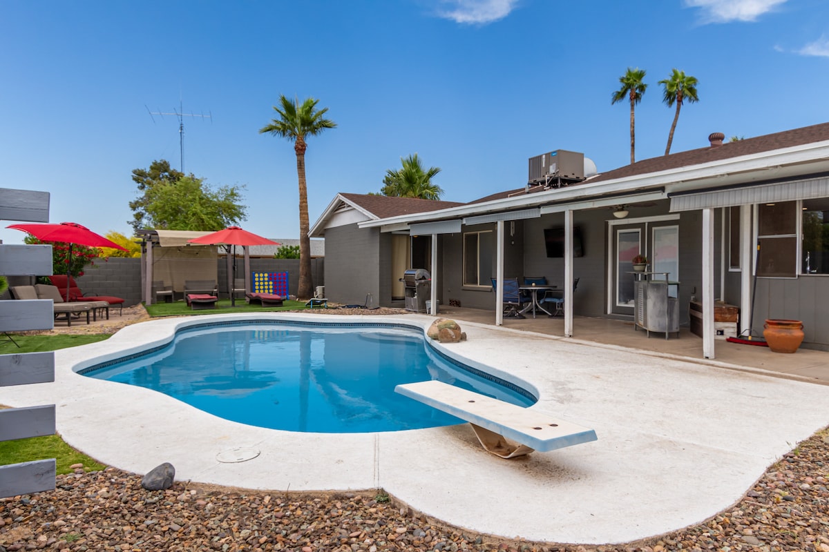 A private backyard features a clear pool surrounded by a spacious concrete patio. Red umbrellas provide shade over lounge chairs. A diving board extends into the water, and a covered seating area is visible. The home is set against a backdrop of palm trees and blue skies.