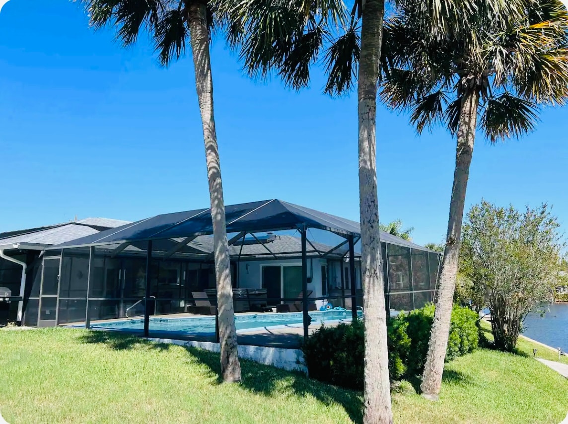 A spacious lanai features a heated pool surrounded by a screened enclosure, providing a bright and airy atmosphere. Lush green grass and palm trees enhance the serene outdoor setting, with views of the nearby saltwater canal visible in the background.