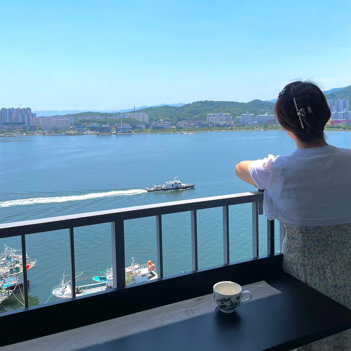 A balcony view showcases a serene expanse of ocean water, gently reflecting the sky. A boat glides across the water, while the coastline and mountains are visible in the distance. A table holds a cup, inviting moments of relaxation amid this scenic backdrop.