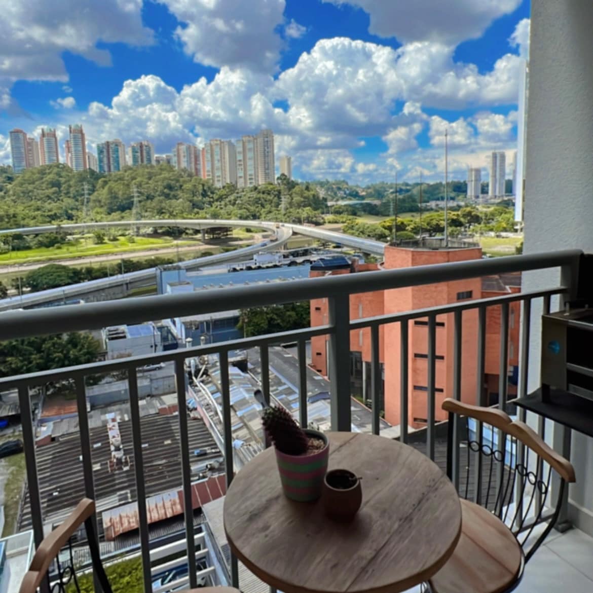 The balcony features a small round table with two chairs and a potted plant. A scenic view of the cityscape and lush greenery is visible in the background, complemented by a bright blue sky with fluffy white clouds.