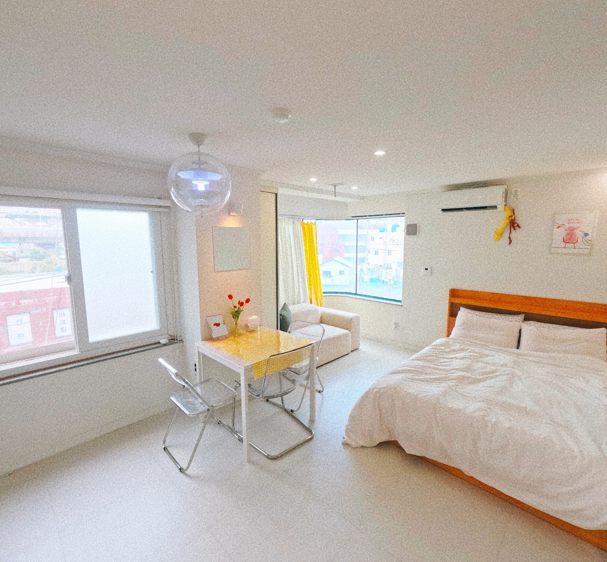 A bright studio space features a spacious bed with white bedding. A glass dining table is paired with two folding chairs, and a soft sofa is positioned near a large window that allows natural light to fill the room. Yellow curtains add a touch of warmth.