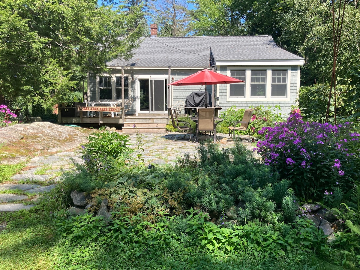 A cottage is framed by lush gardens, featuring a stone patio with a table shaded by a red umbrella. Seating is arranged for outdoor dining, while vibrant flowers bloom nearby. The large windows suggest a well-lit interior, enhancing the natural setting.