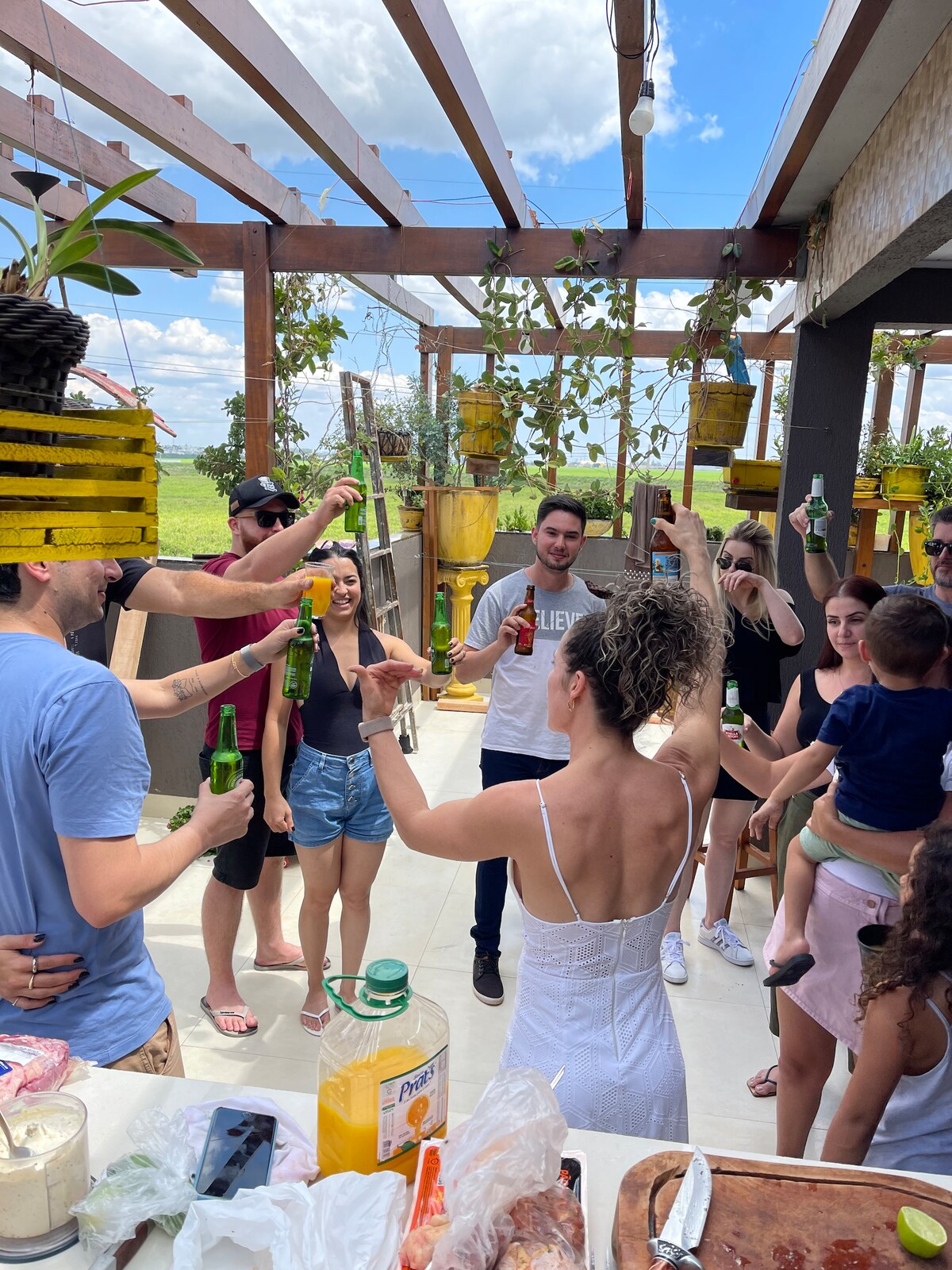A rooftop space is filled with people celebrating while holding drinks. The area features wooden beams, potted plants, and a scenic view in the background. Natural light from above creates an inviting atmosphere, and a table nearby displays food and beverages.
