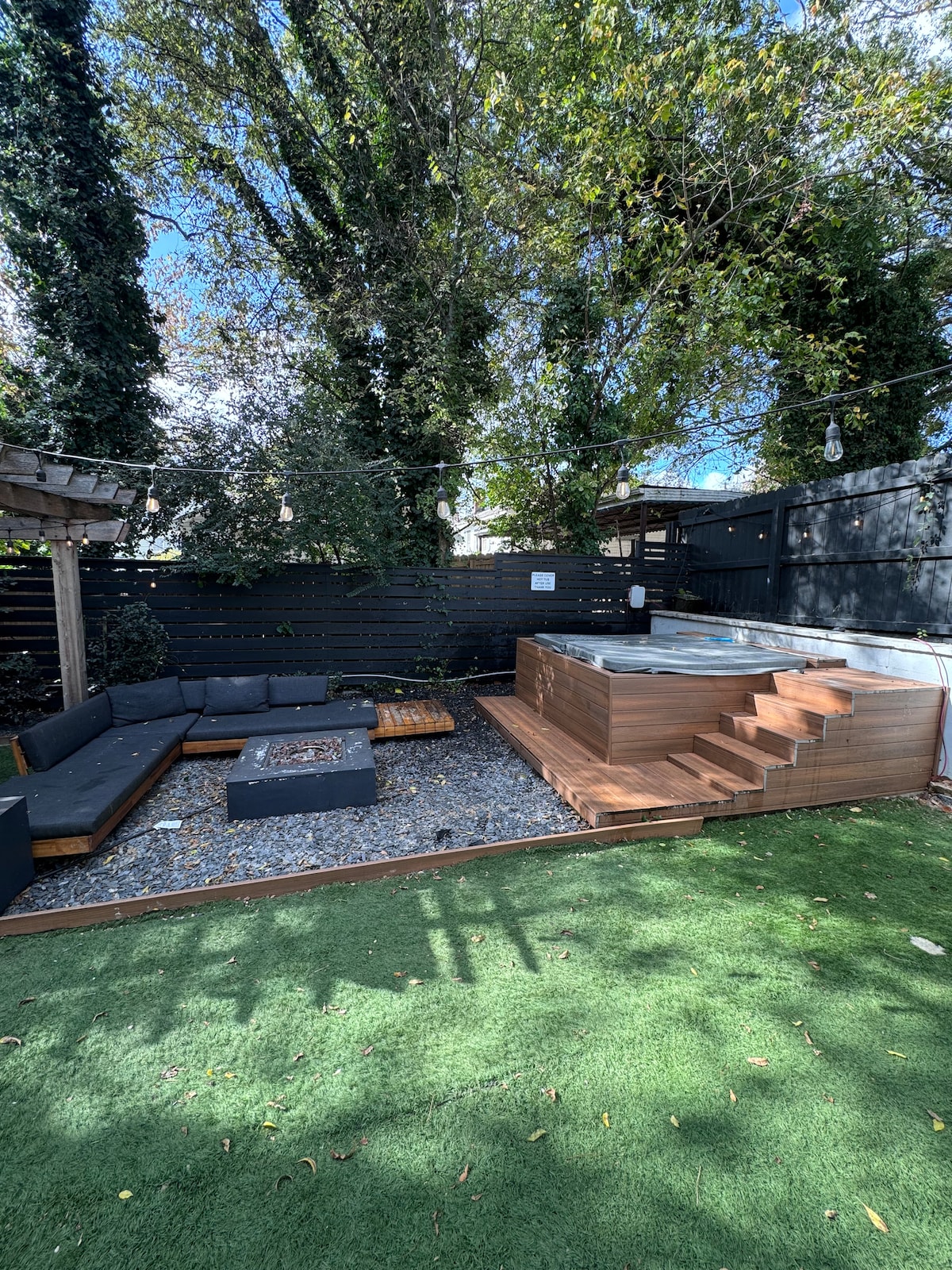 An outdoor relaxation area features a wooden hot tub surrounded by lush greenery. Comfortable seating is arranged around a fire pit on gravel, with a well-maintained lawn providing a soft contrast. Natural light filters through the overhead tree canopy.