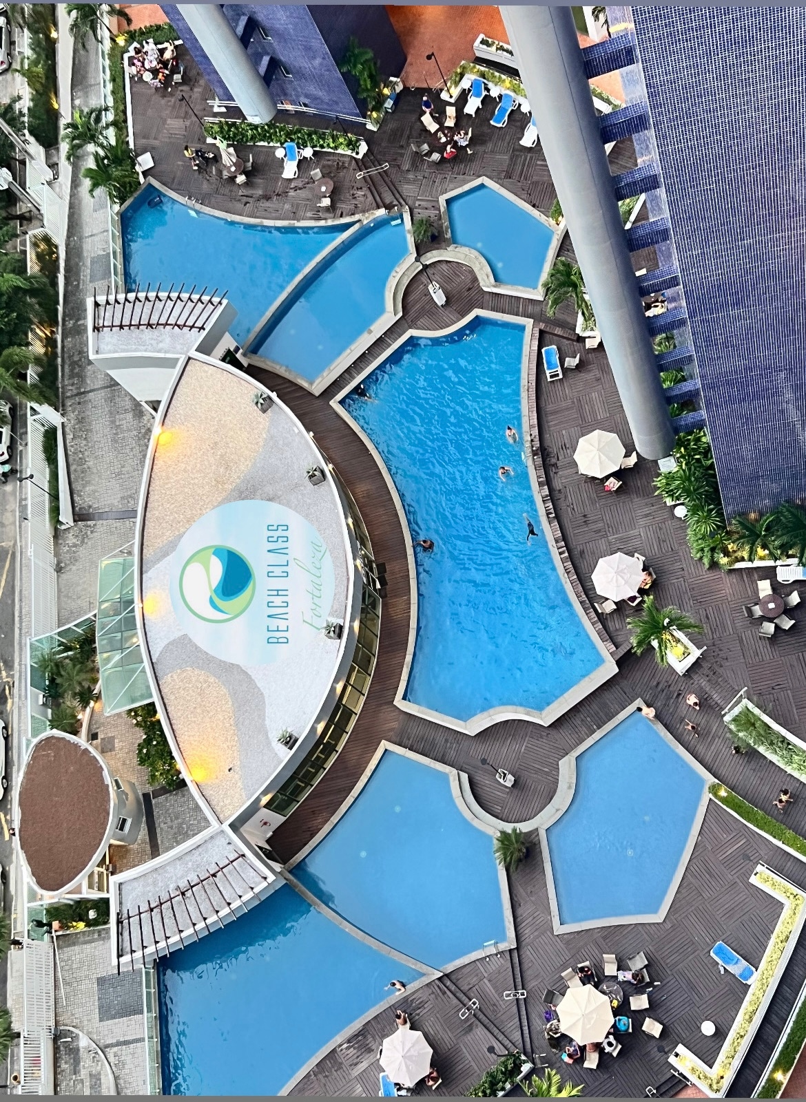 Aerial view of multiple swimming pools showcasing varying shapes and sizes, surrounded by spacious sun decks dotted with umbrellas and lounge chairs. The area is framed by well-maintained landscaping, conveying a resort-like atmosphere.