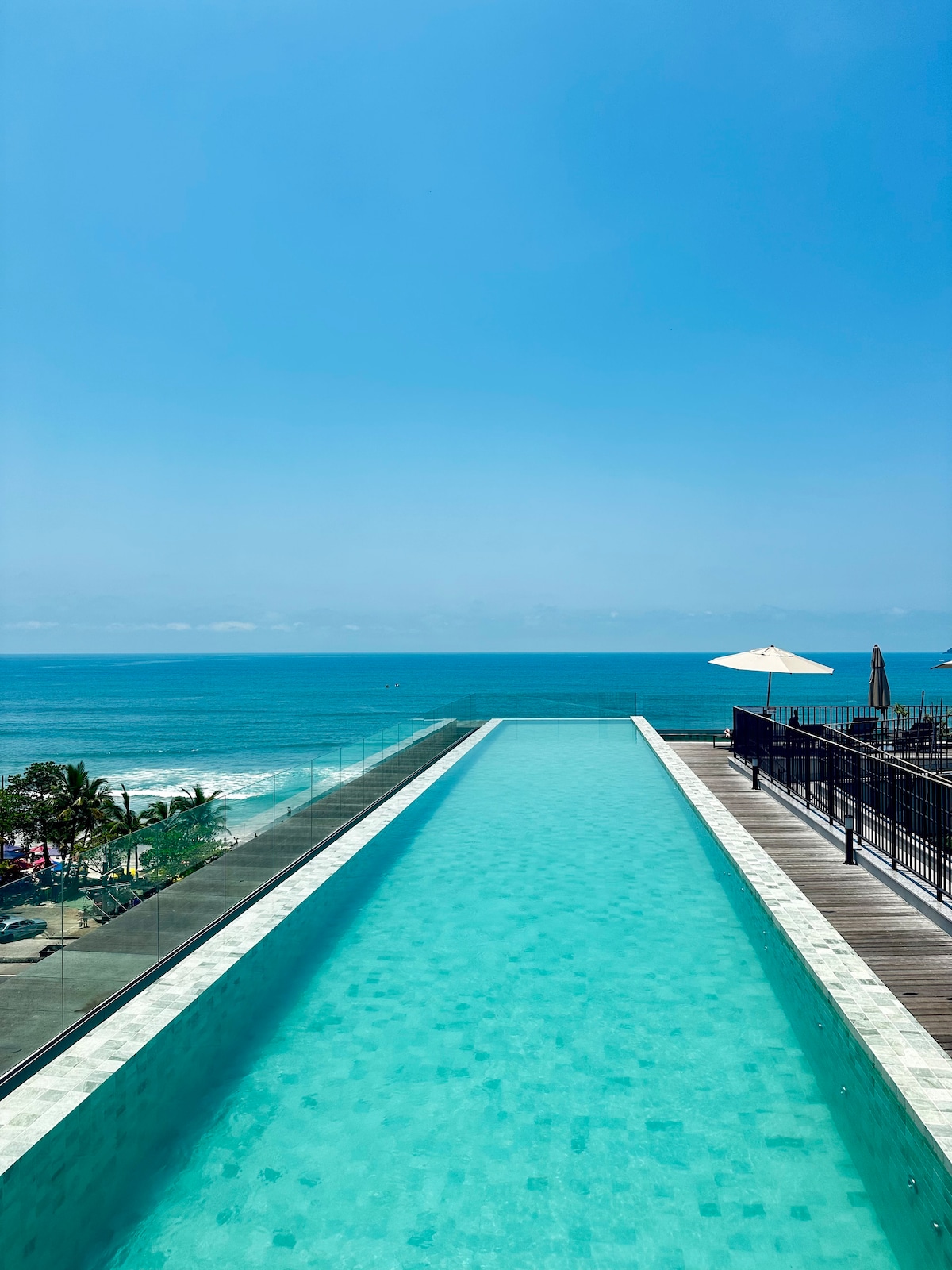 An expansive infinity pool extends towards the horizon, blending seamlessly with the ocean. The vibrant turquoise water reflects the clear blue sky, creating a tranquil atmosphere. Lounge chairs and umbrellas are positioned along the poolside, offering relaxation spots with stunning views of the beach.