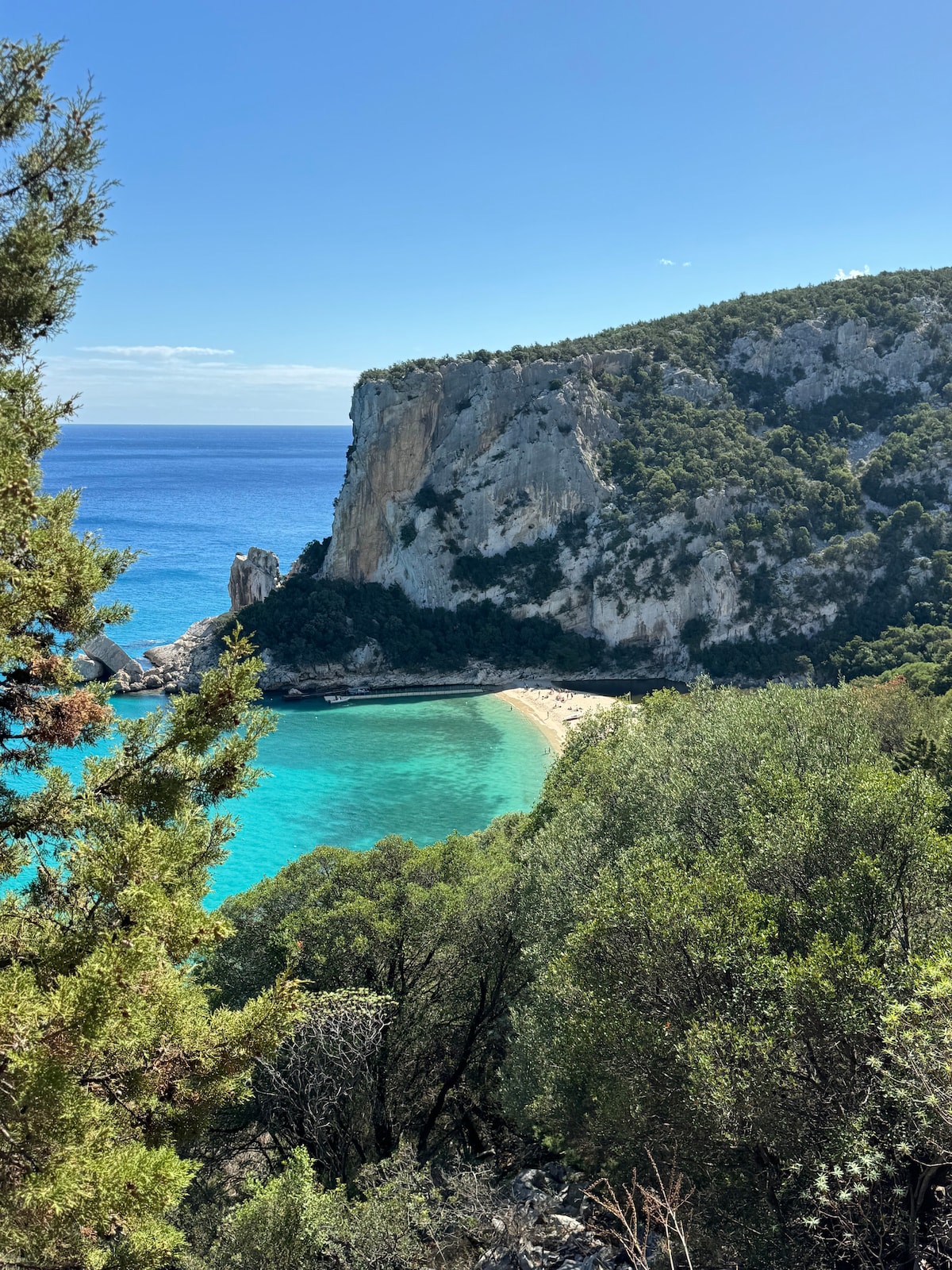 A secluded beach is nestled between rocky cliffs, surrounded by lush greenery. The turquoise waters gently lap at the sandy shore, creating a calm and serene atmosphere. The sunlight reflects off the water, enhancing the natural beauty of the landscape.