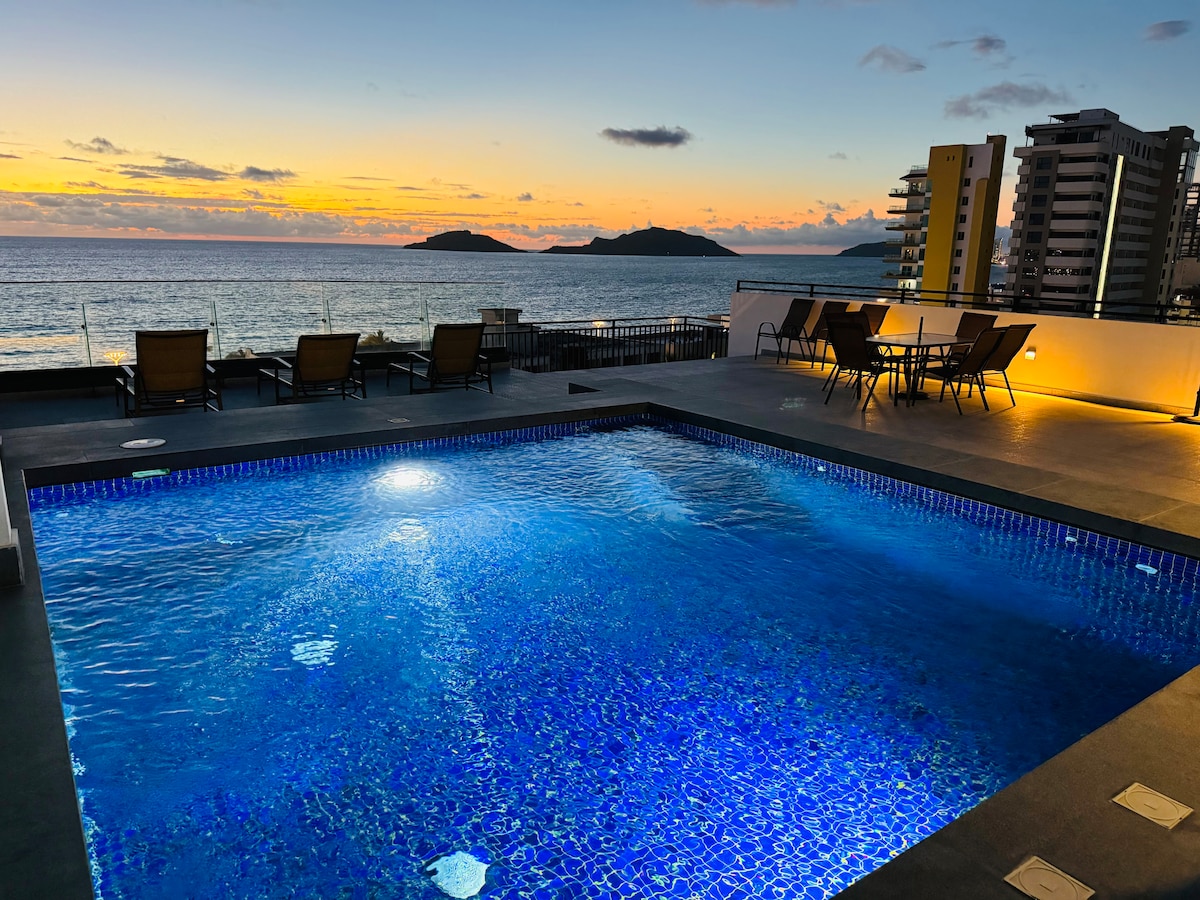 A modern pool area features a vibrant blue swimming pool with sparkling water under a sunset sky. Lounge chairs provide seating along the pool's edge, while a scenic view of the ocean and islands is visible in the background.