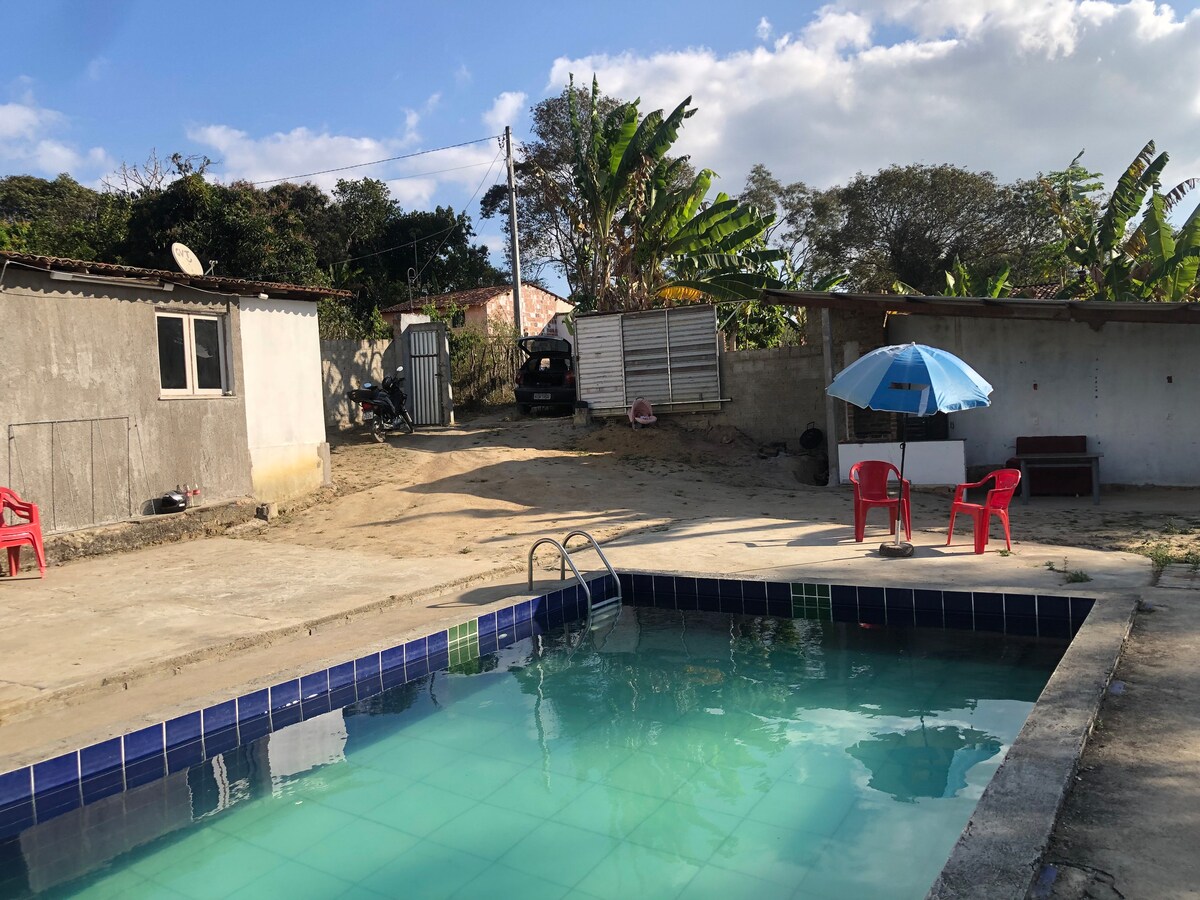 The pool area features a clear blue pool surrounded by a concrete patio. Two red chairs and a blue umbrella provide shaded seating. The background includes a simple structure with a window and a grassy area, along with several trees bringing a natural touch.