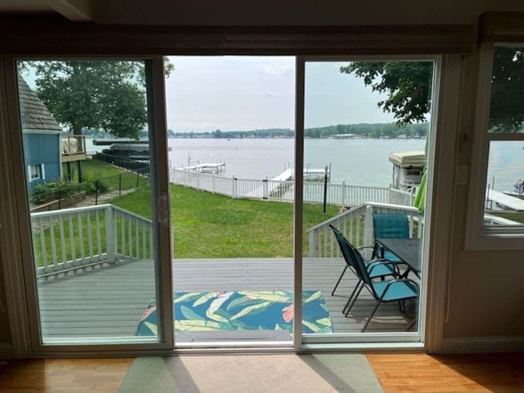 A glass sliding door reveals a spacious deck with a colorful rug and outdoor seating. The view showcases a peaceful lakeside landscape, with green grass leading to the water where boats are docked in the distance.