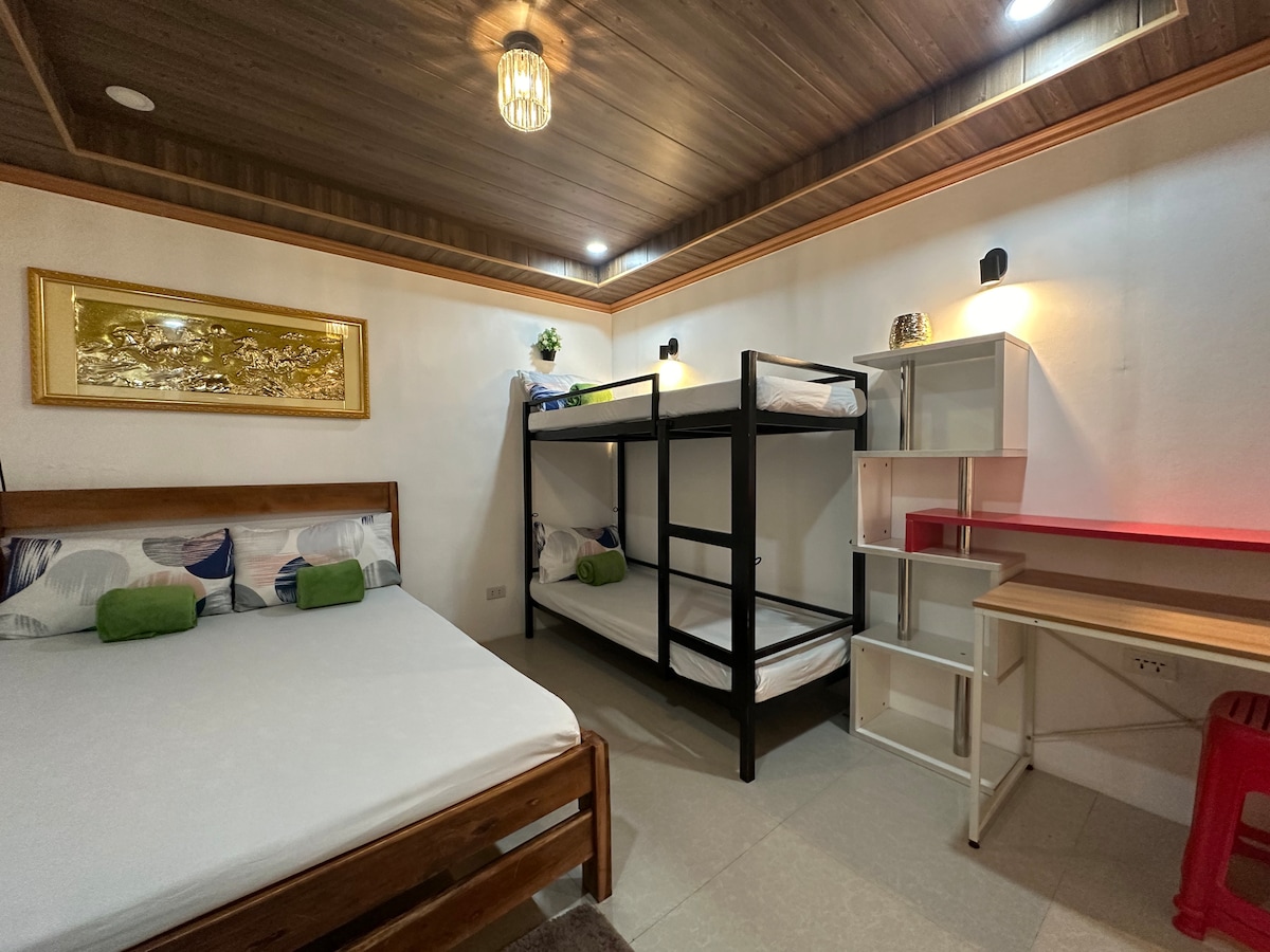 A cozy studio room is featured, showcasing a spacious bed alongside two bunk beds. A simple writing desk and shelf are included, while wall-mounted lights provide illumination. Green accents appear on the bedding, and a decorative piece is mounted above the bed.