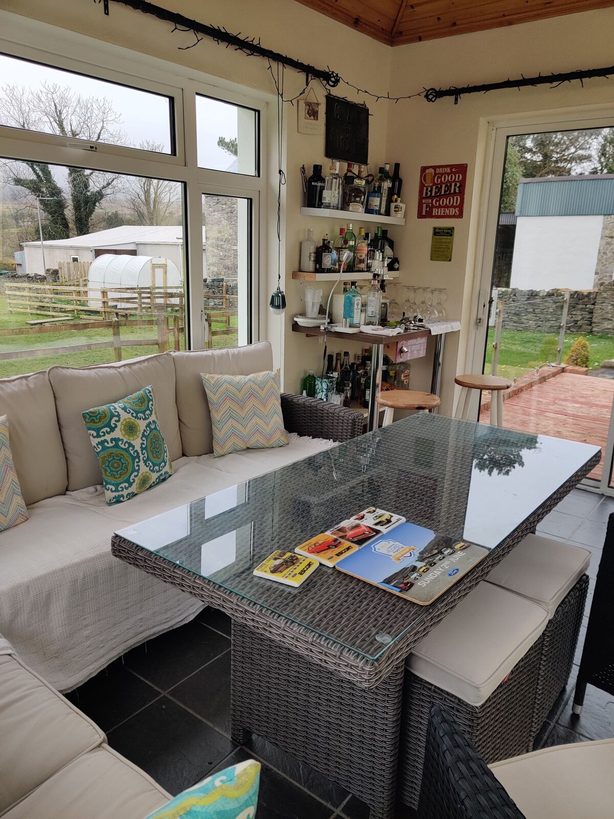 A bright sunroom features a comfortable seating area with cushions in various colors. A glass-topped table sits at the center, surrounded by four wicker chairs. Shelving displays a variety of beverages and accessories. Large windows provide views of the green garden outside.
