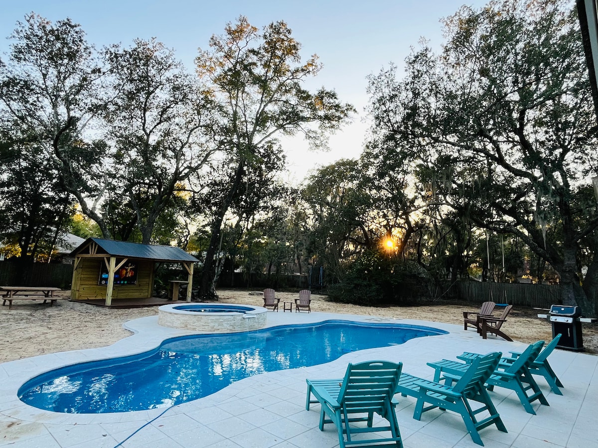 An outdoor area features a large pool with a jacuzzi, surrounded by inviting turquoise lounge chairs. Sunlight filters through the trees, highlighting a gazebo nearby. A sandy area includes picnic tables, providing space for leisurely outdoor gatherings.