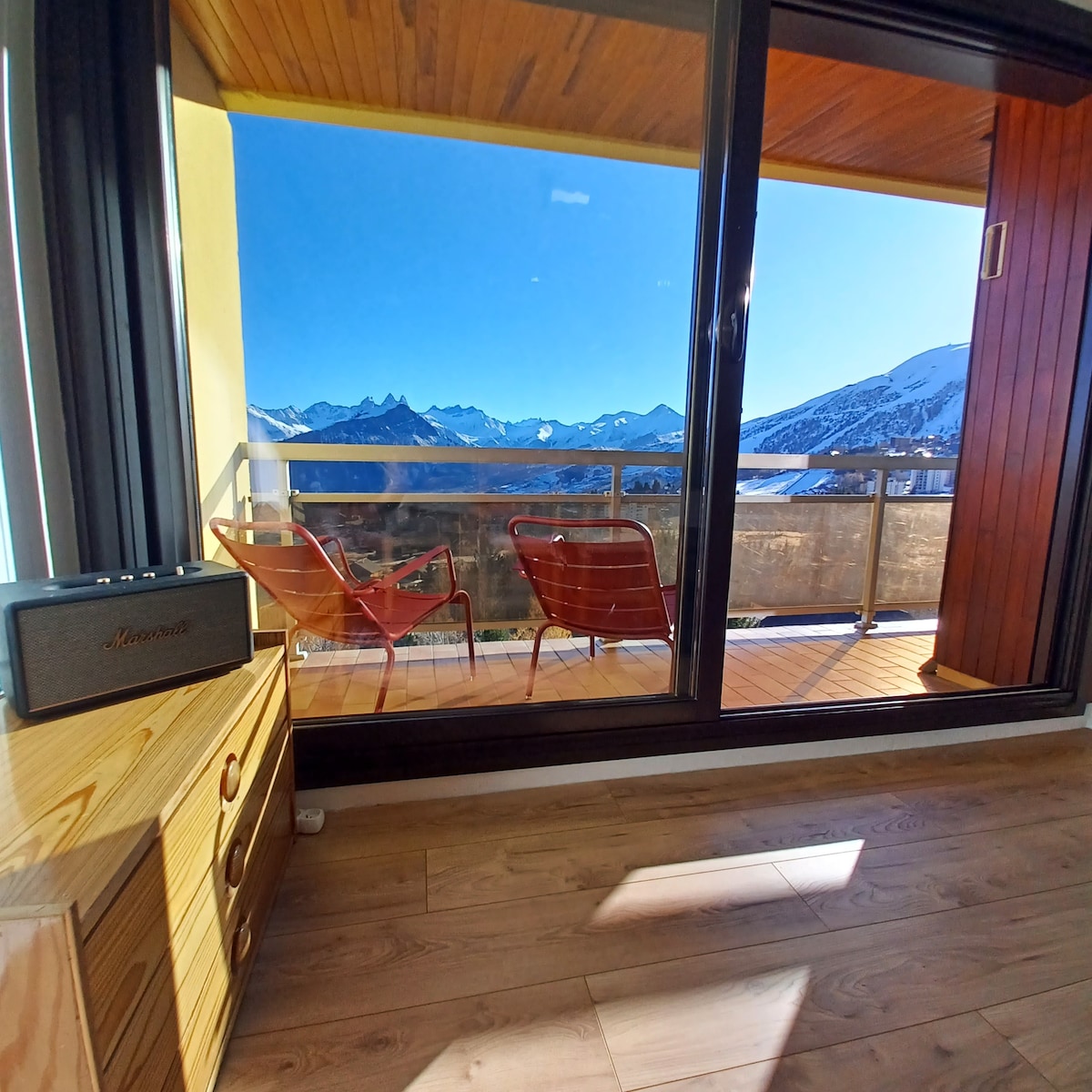 A balcony is featured with two chairs facing panoramic mountain views. The space is accessible through large glass doors, allowing natural light to illuminate the interior. A wooden dresser is positioned beside a vintage speaker, adding functionality and character to the area.