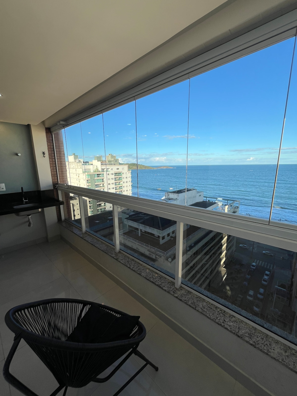 A balcony features expansive glass panels overlooking the ocean. The serene view includes the shoreline, with buildings positioned nearby. An accent chair is placed on the balcony, adding to the space's usability while promoting a connection with the outdoor environment.