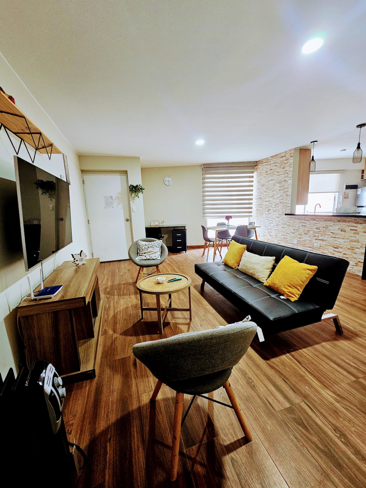 A modern living area features a comfortable sofa bed adorned with yellow and gray cushions. A wooden coffee table sits in front, while a wall-mounted television provides entertainment. Natural light enters through the window treatments, illuminating the warm wood flooring and stylish decor.