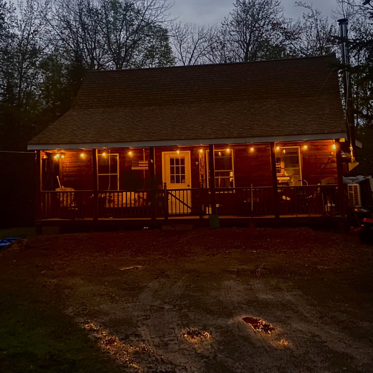 The cabin exterior is softly illuminated by warm string lights, creating a welcoming glow. The front porch, furnished with seating, provides easy access through a central door. Surrounding trees are partially visible in the evening setting, complementing the rustic charm of the cabin.