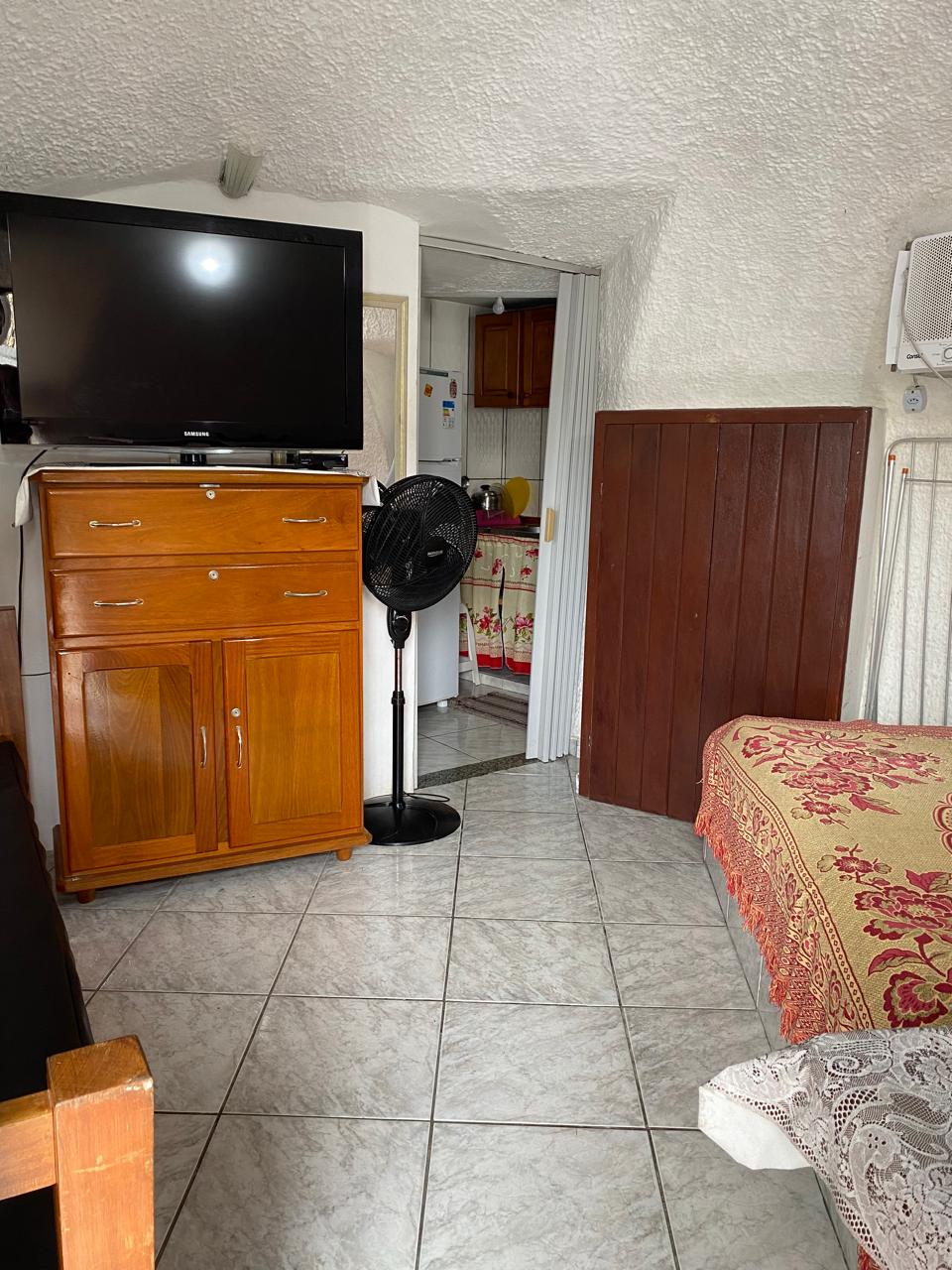 A compact living space features a wooden dresser with multiple drawers, a large television mounted on the wall, and a fan on a stand. A cozy bed adorned with patterned bedding is positioned in the corner, with tiled flooring reflecting the room's light.