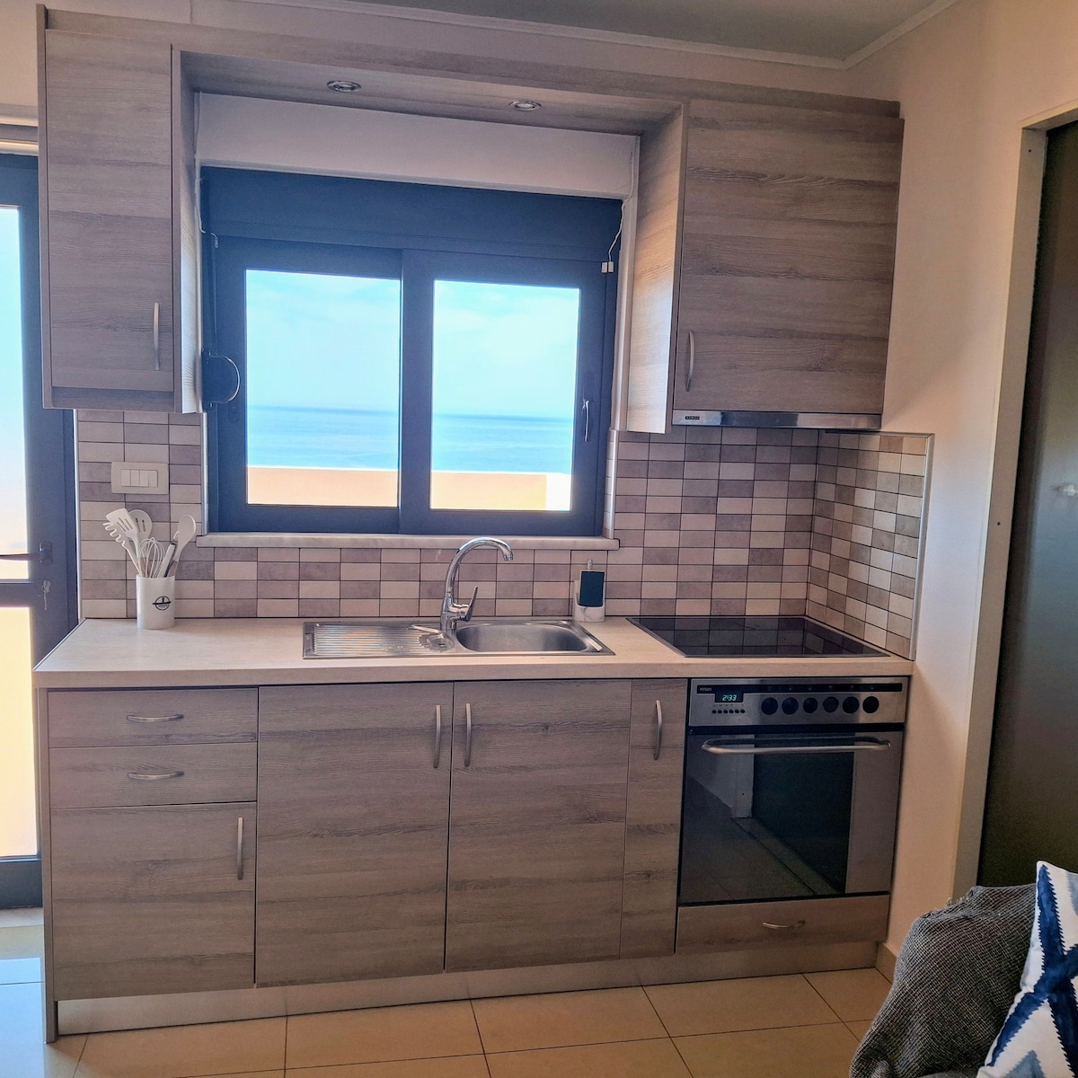 A modern kitchen is visible, featuring light wood cabinets with sleek finishes. A window offers an unobstructed view of the sea. Included are a stainless steel sink, a stove, and an oven, all arranged against a backsplash of decorative tiles.