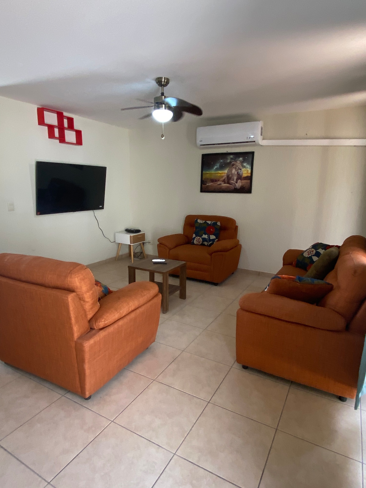 A comfortable seating area is arranged with two armchairs and a sofa, all featuring orange upholstery. A small table is positioned at the center, with a flat-screen television mounted on the wall opposite. The space is illuminated by a ceiling fan and a decorative wall piece.