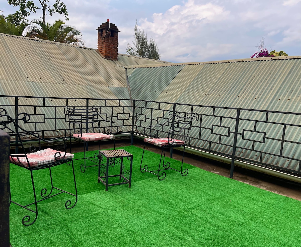 A terrace area features artificial grass, creating a serene outdoor moment. Metal chairs and a small table are arranged for relaxation, with a sloped roof adding character to the space. The surrounding greenery offers a touch of nature, complemented by atmospheric clouds overhead.