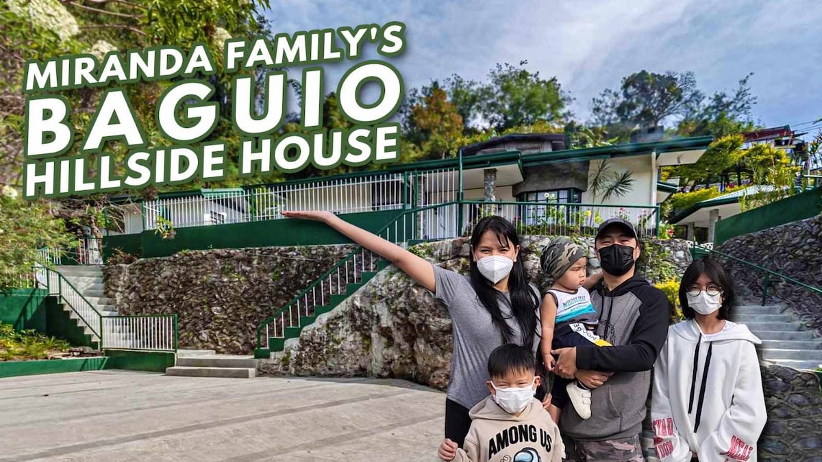 A family stands in front of Baguio Hillside House, with a welcoming patio area featuring stone walls and green railings. Surrounding trees provide a natural backdrop, enhancing the inviting setting. The house itself is visible in the background, characterized by a layered architectural design.