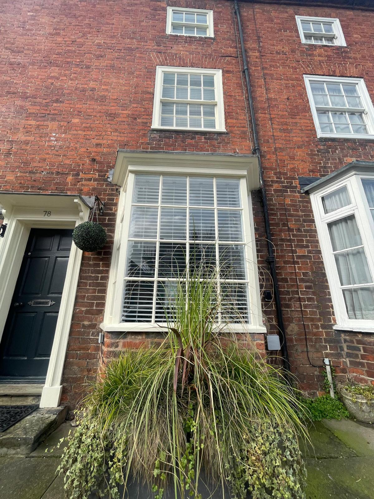 The exterior of a Georgian townhouse is shown, featuring red brick walls and multiple sash windows. A decorative window box with plants is visible below the main window, and a door with a dark finish stands at the entrance. Attention is drawn to the period architectural details.