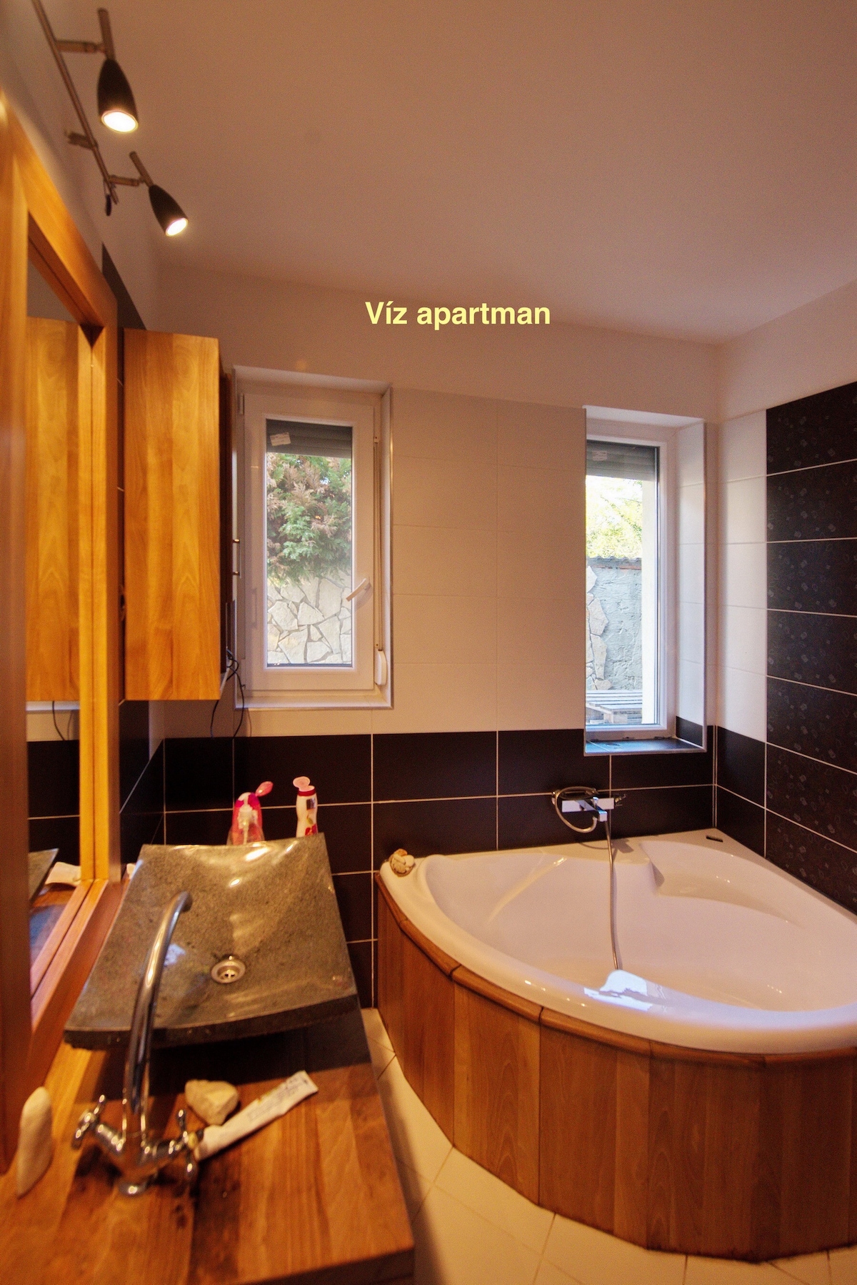 The bathroom features a corner bathtub with a wooden surround. A modern sink is positioned next to a large mirror, and natural light enters through two small windows, highlighting the contrasting wall tiles in dark and light shades.