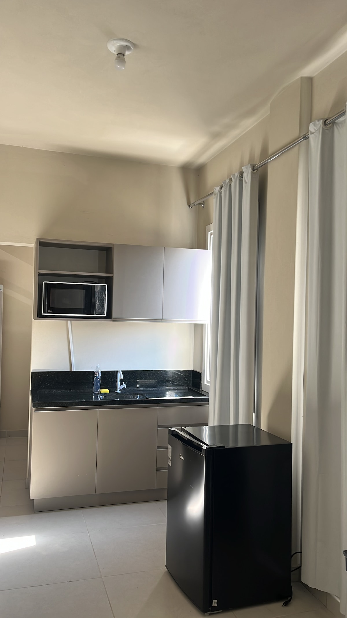 A compact kitchen area features modern cabinetry and a sleek black fridge. A microwave is positioned on the countertop, alongside a sink. Light-colored walls and large windows allow natural light to fill the space, complemented by flowing white curtains.