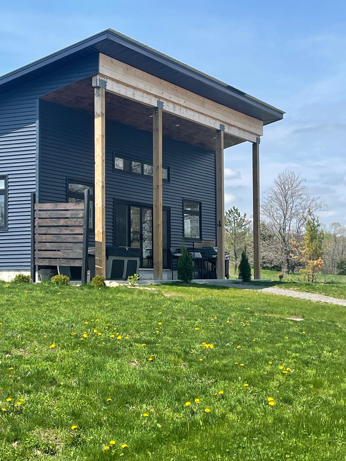 The exterior of the cottage is showcased with a modern, slatted porch that offers shade. A well-maintained lawn features patches of wildflowers, while large windows reflect natural light. The structure is painted in a deep blue hue, harmonizing with the surrounding greenery.