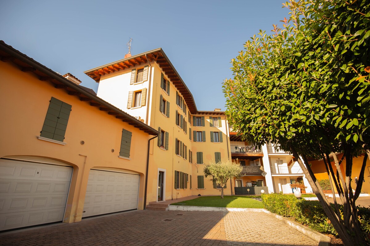 An exterior view of a charming building featuring warm yellow walls and classic green shutters. The structure displays multiple floors with balconies overlooking a well-kept garden. A paved area provides access to garages, surrounded by neatly trimmed hedges and a small tree.