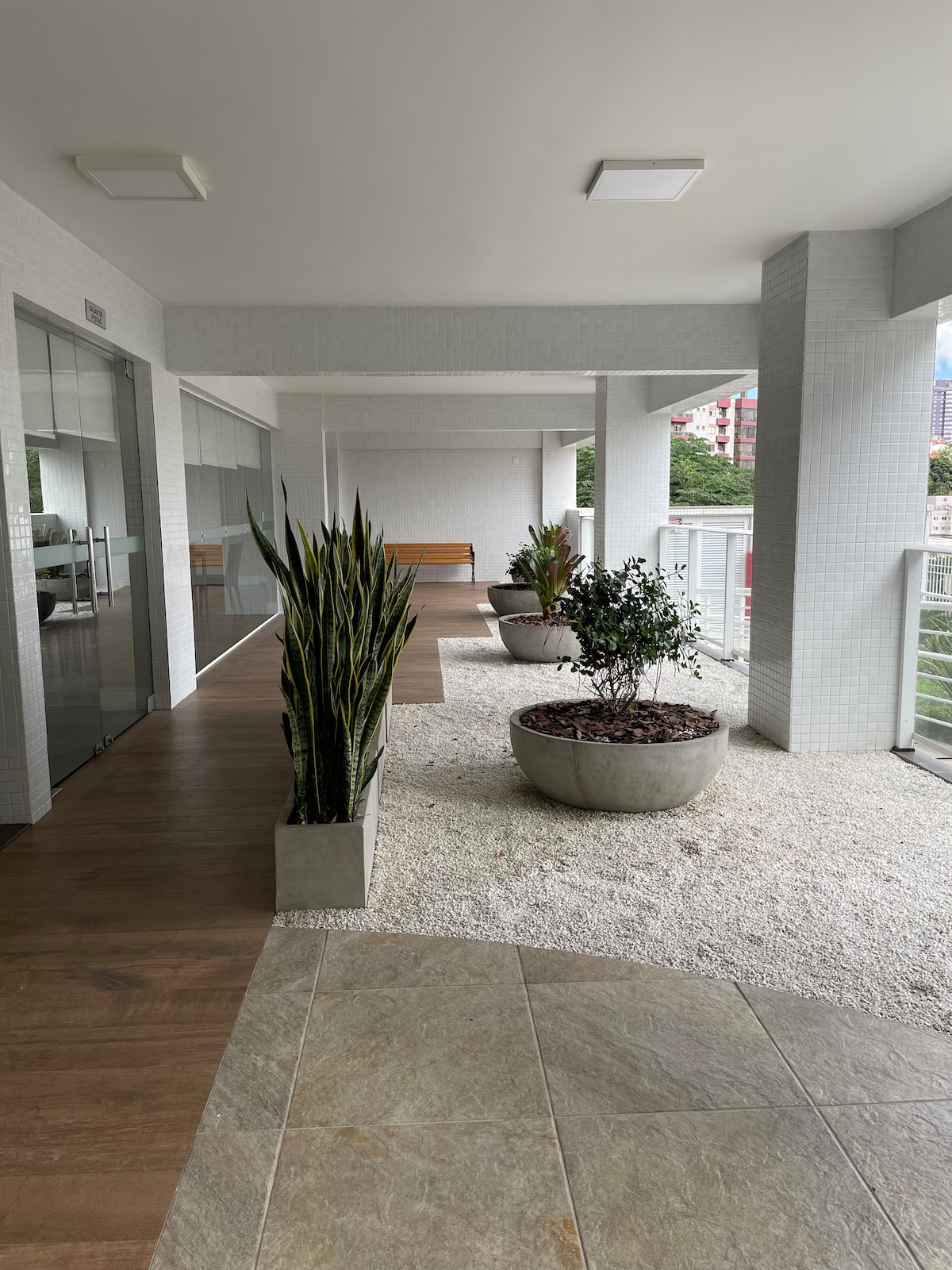 A serene corridor is lined with modern planters filled with greenery, creating a refreshing atmosphere. The space is brightened by natural light filtering through large glass doors, with sleek flooring extending throughout.