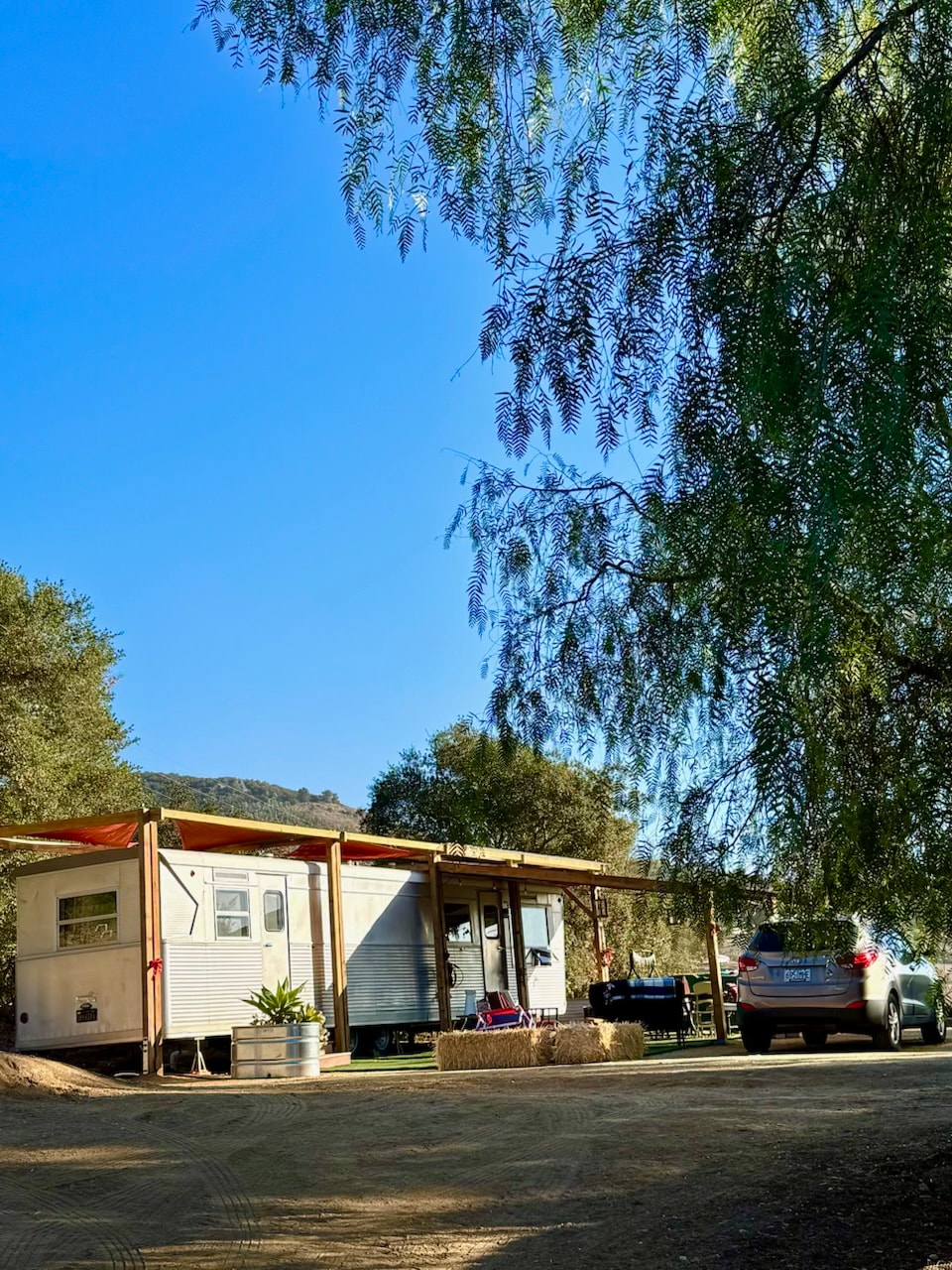 A vintage travel trailer is positioned under a shaded awning, surrounded by greenery. A vehicle is parked nearby, with a clear blue sky above and gentle trees framing the scene. The outdoor space is prepared for relaxation and enjoyment.