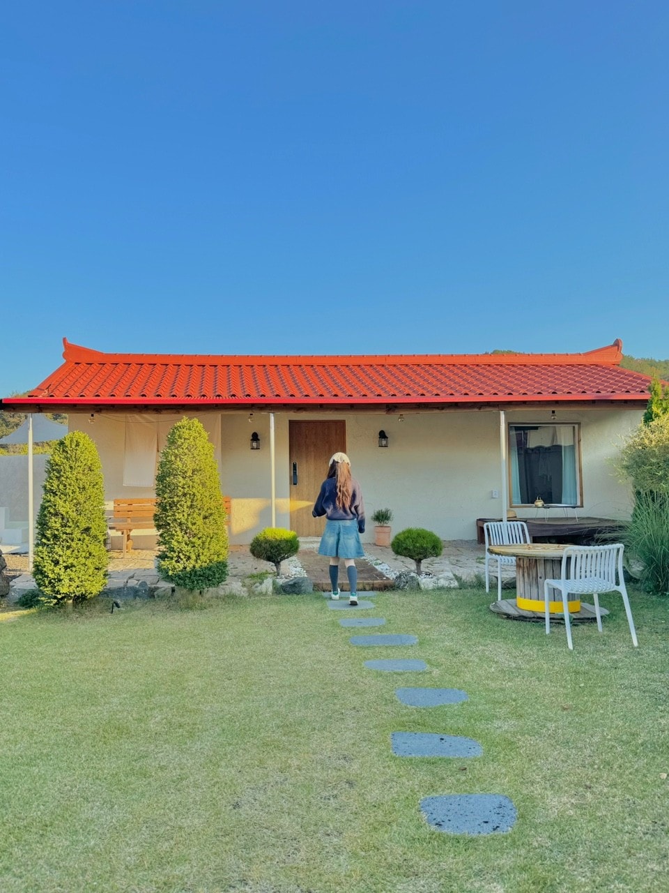 A charming home with a red roof is nestled among greenery. A pathway of stepping stones leads to the entrance. Flanking the house are neatly trimmed shrubs, while outdoor seating is positioned beside a table on the lawn, inviting relaxation in the serene surroundings.