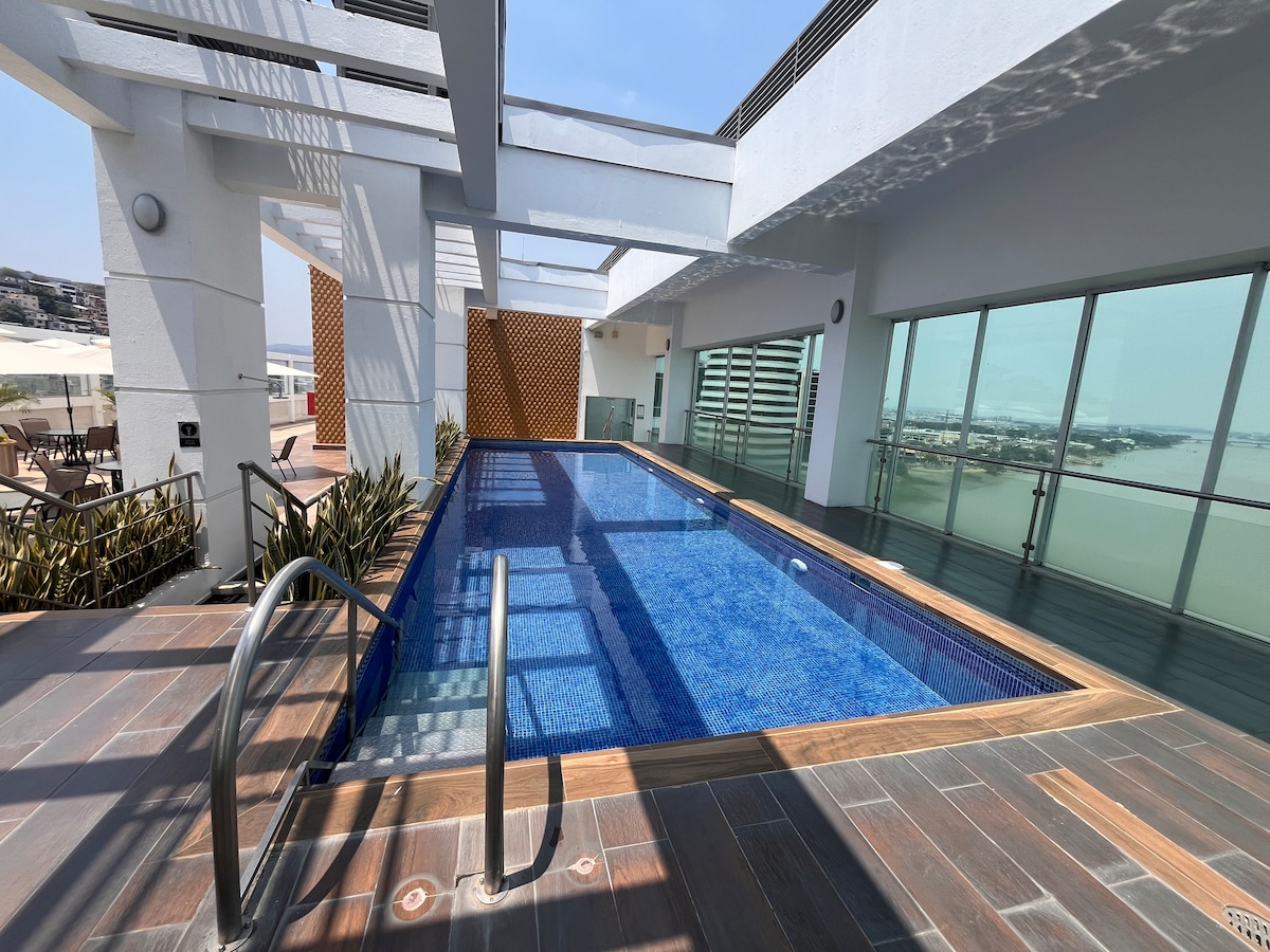 A rooftop pool with clear blue water is surrounded by a sleek wood deck and modern railing. Large windows provide an expansive view of the surroundings, while sheltered areas feature stylish design elements and greenery, creating a serene retreat.