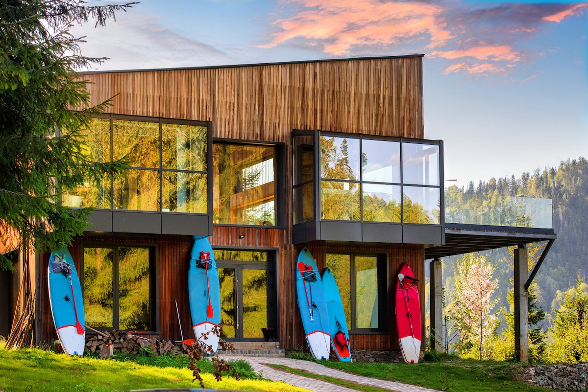 The modern chalet features a striking wooden exterior with large glass windows reflecting the surrounding landscape. Colorful paddleboards are displayed on the front, while a pathway leads to the entrance, framed by lush greenery and wooded hills in the background.