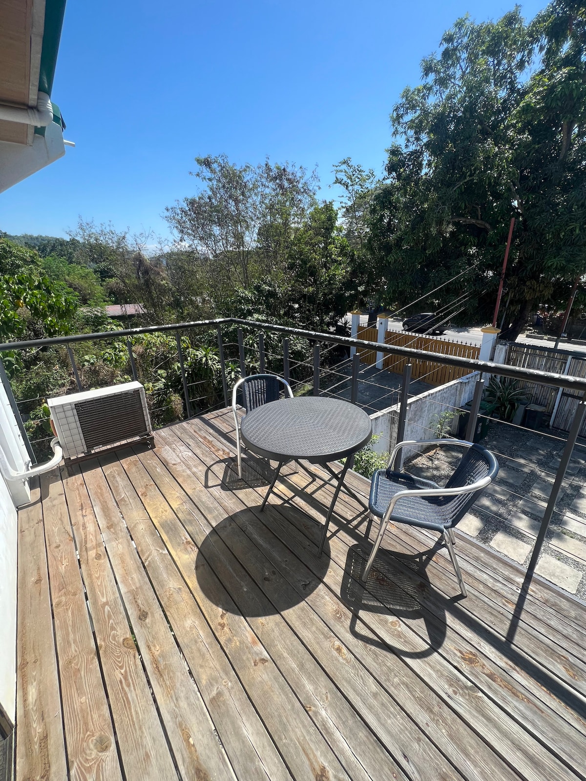 A wooden deck area is shown, featuring a round table surrounded by two chairs. Lush greenery can be seen in the background, adding to the outdoor ambiance. Clear skies and sunlight illuminate the space, creating an inviting spot for relaxation.