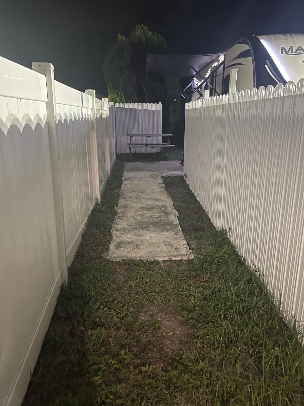 A narrow pathway lined with white fencing is seen, leading to a small picnic table positioned at the end. The grass borders the sides of the concrete walkway, which is illuminated by soft night light, creating a serene outdoor space.