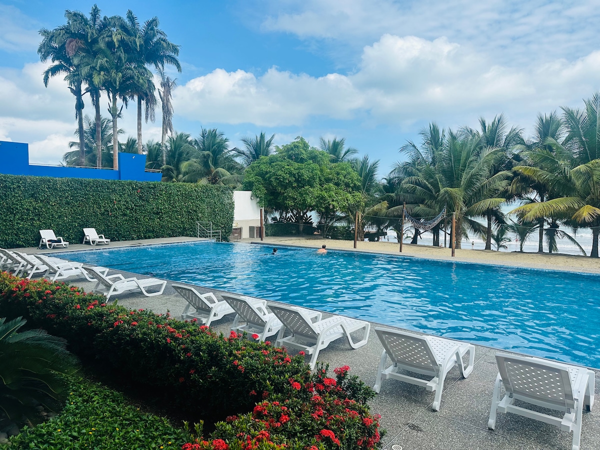 A clear swimming pool is surrounded by lush tropical palm trees and vibrant greenery. Lounge chairs are positioned along the poolside, offering ample seating. The calm waters reflect the blue sky above, creating a serene environment for relaxation.