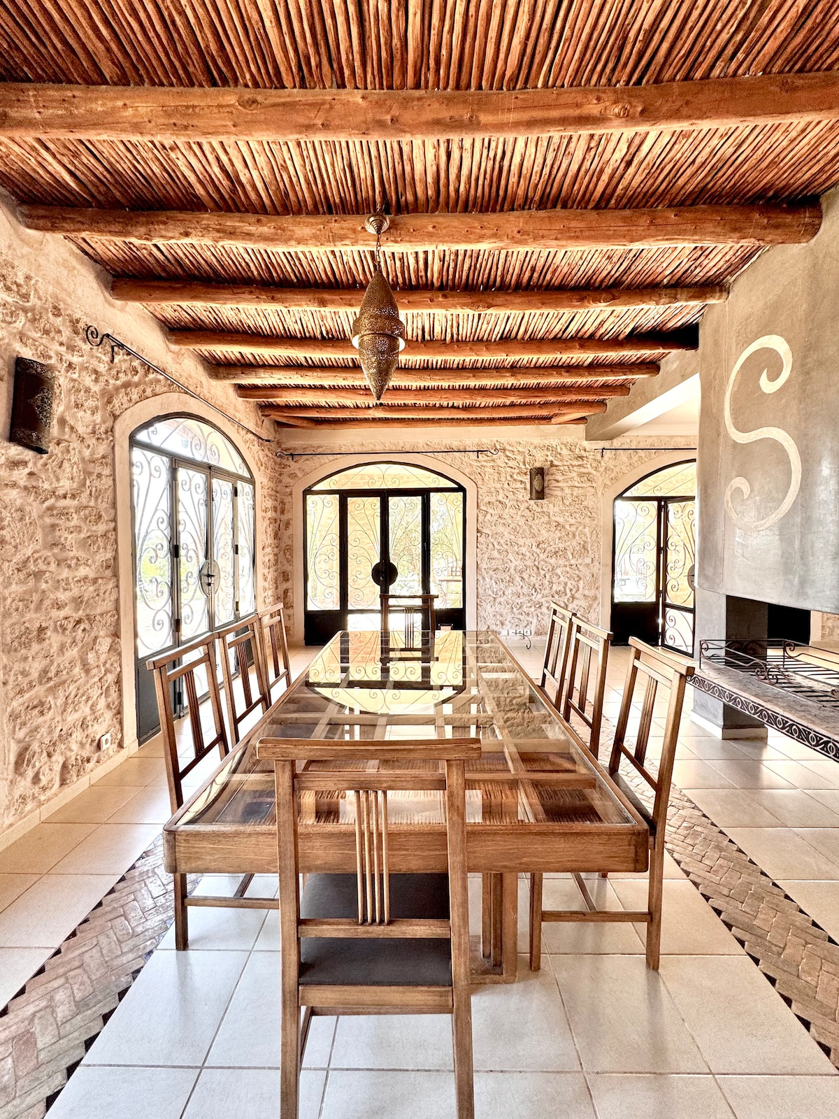 A spacious dining area is highlighted by a large glass-top table surrounded by wooden chairs. Exposed wooden beams form the ceiling, complementing the textured stone walls. Large arched doors allow natural light to fill the space, enhancing the inviting atmosphere.