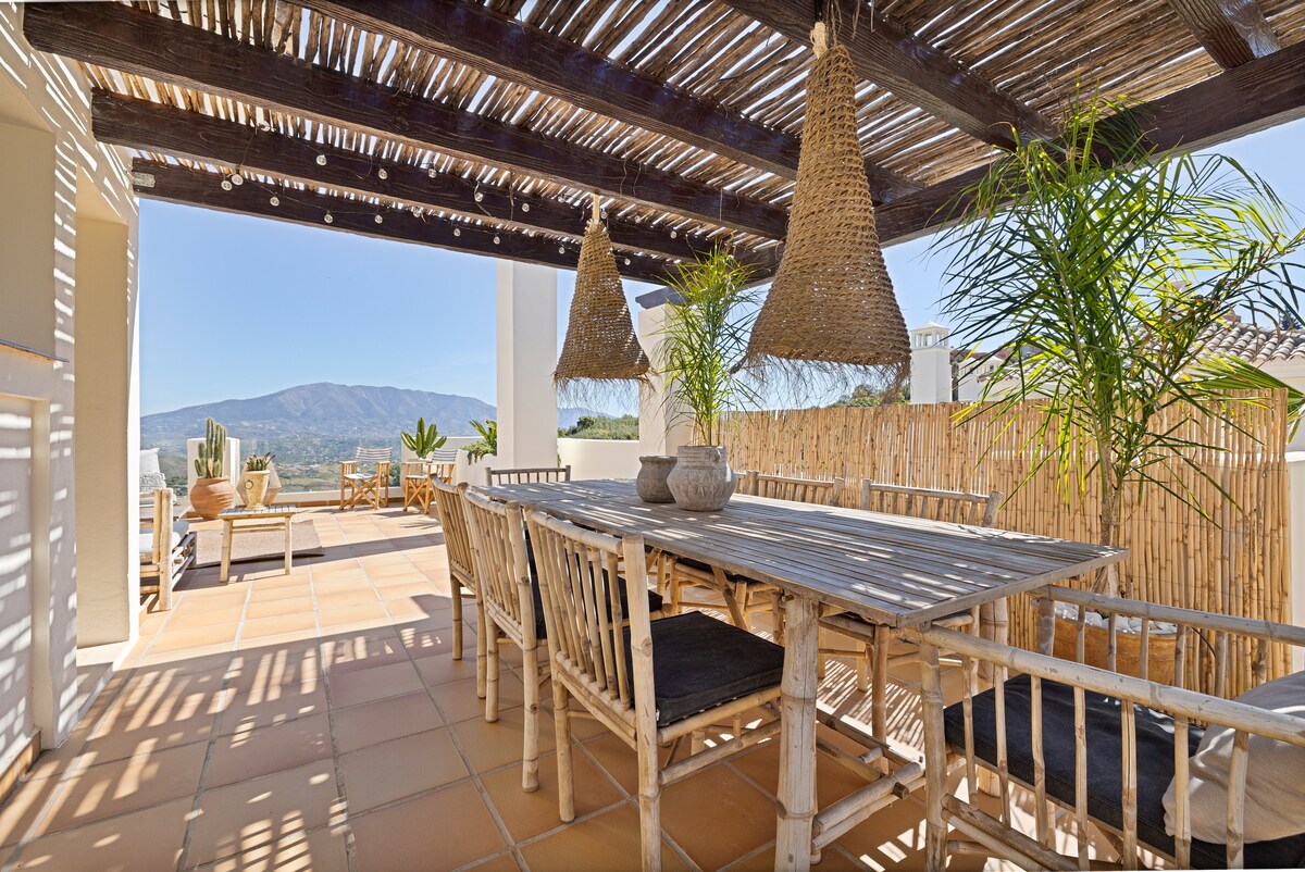 A dining area is presented with a wooden table surrounded by bamboo chairs, shaded by a thatched roof. Two woven pendant lights hang above, while potted plants and a mountainous backdrop enhance the serene outdoor setting.